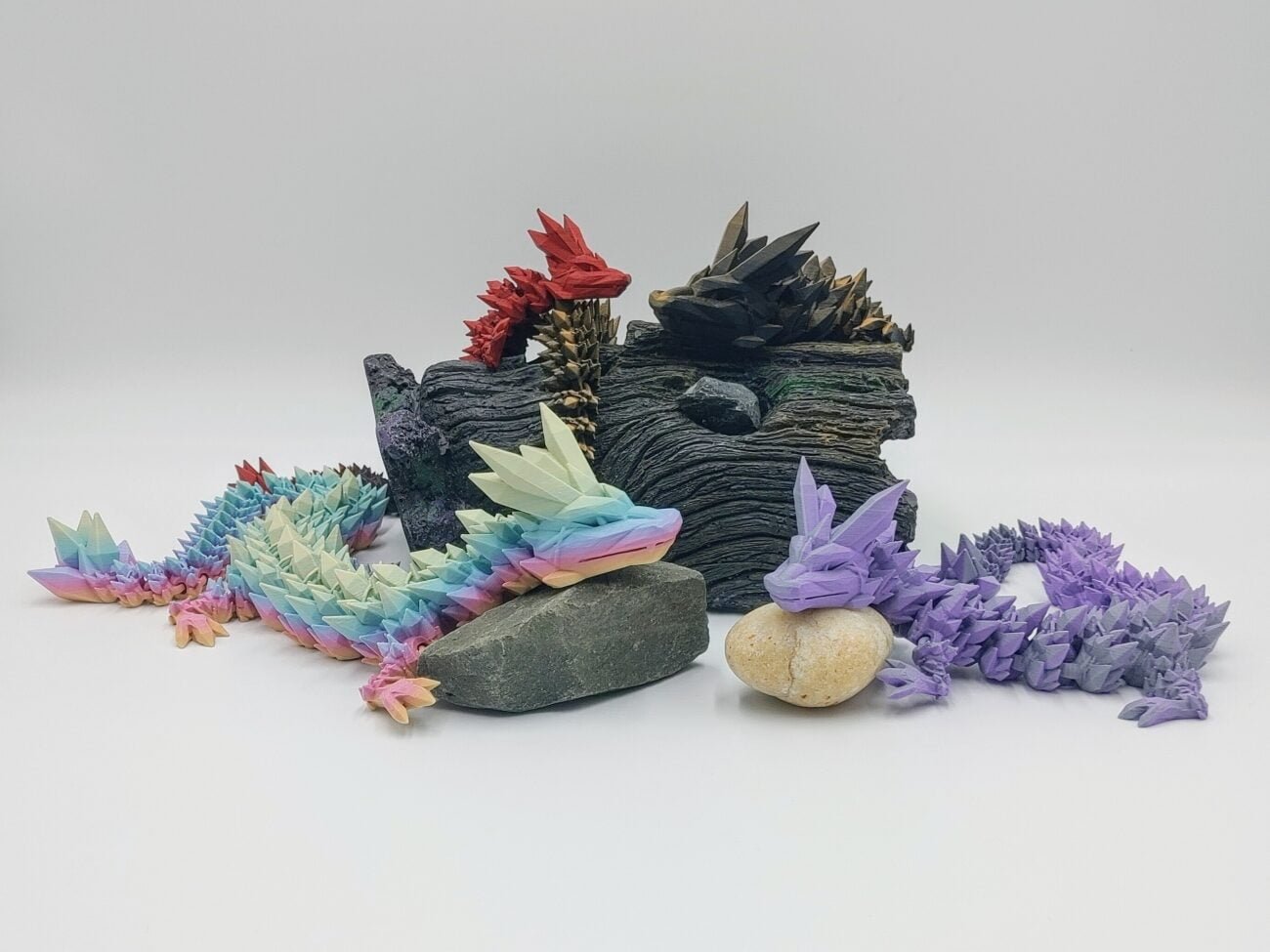 Articulated Crystal Dragons 3D Printed in Matte Dual Tone Hues and Rainbows Articulated Crystal Dragon Sensory Toys in Matte Dual Tone Hues and Rainbows Sitting on Rocks and a Log with a White Background