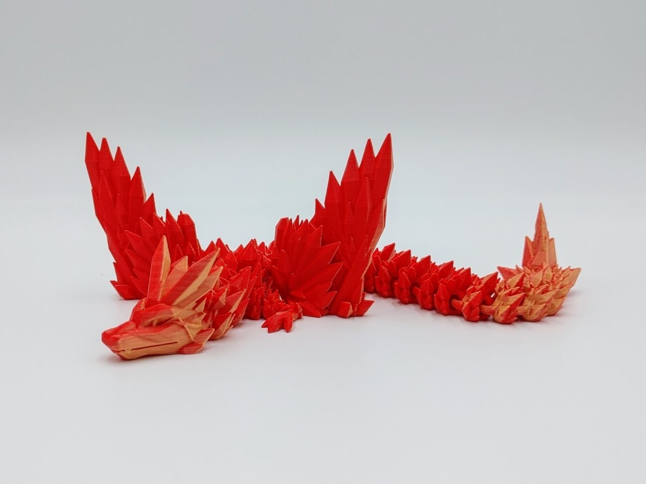 Articulated Crystalwing Dragon 3D Printed in Matte Red to Yellow Articulated Crystalwing Dragon Sensory Toys in Matte Red to Yellow Sitting on Rocks on a White Background