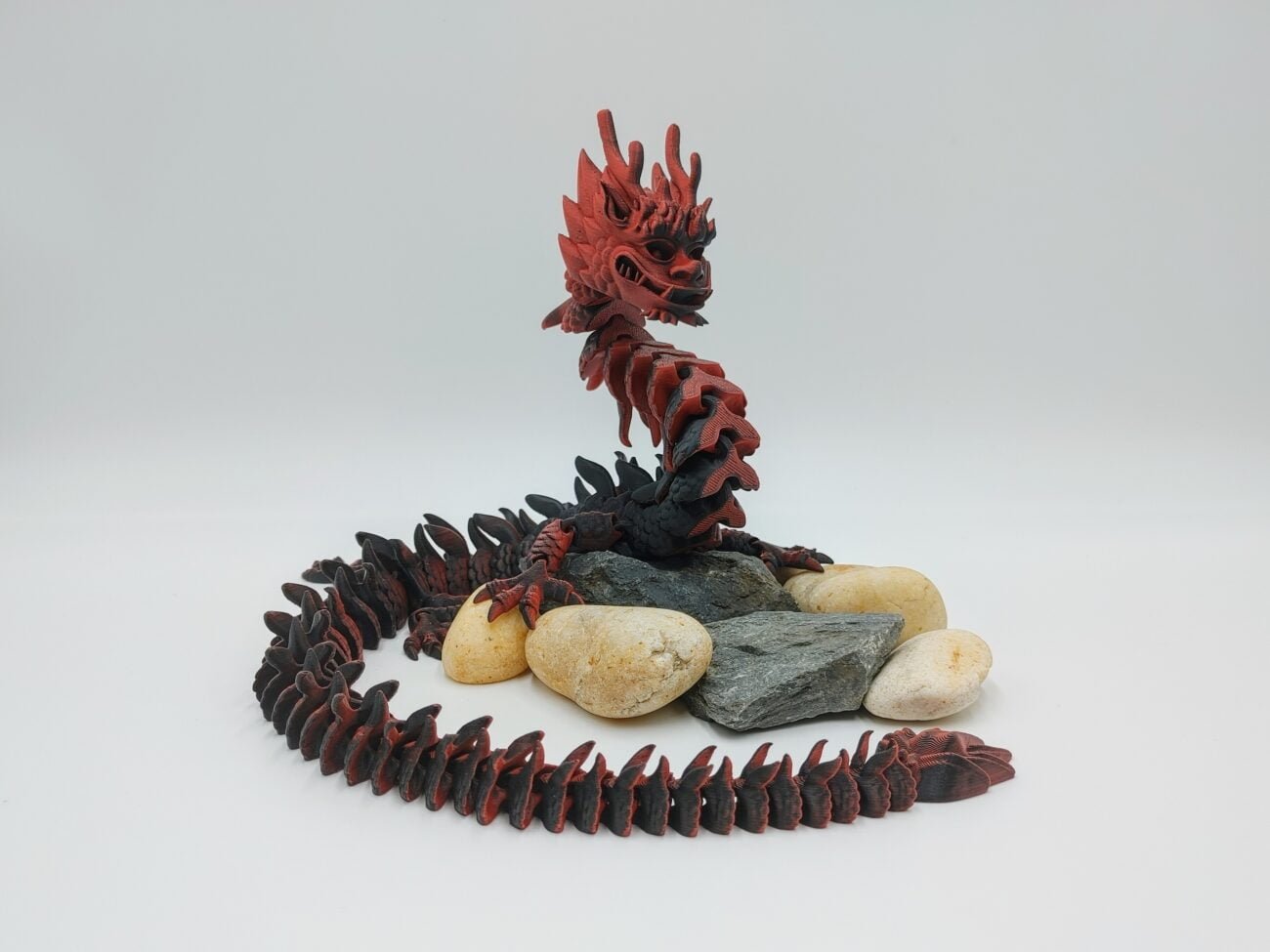Articulated Imperial Dragon 3D Printed in Matte Red to Black Articulated Imperial Dragon Sensory Toys Designed by Flexy Factory in Matte Red to Black Sitting on Rocks with a White Background