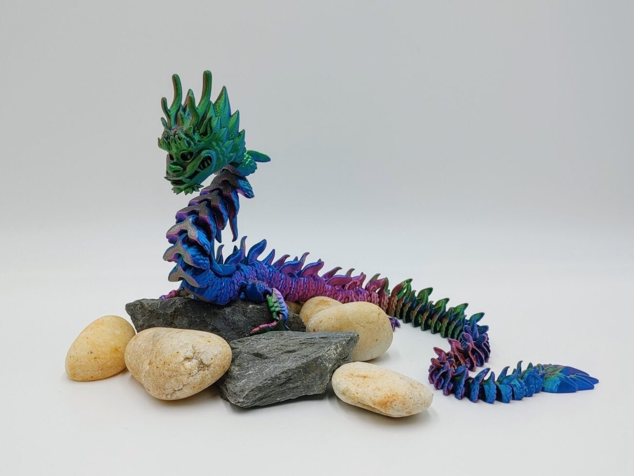 Articulated Imperial Dragon 3D Printed in Silk Tri Colour Red to Green to Blue Articulated Imperial Dragon Sensory Toys Designed by Flexy Factory in Silk Tri Colour Red to Green to Blue Sitting on Rocks with a White Background